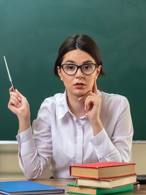 thinking putting finger on cheek young female teacher wearing glasses points at blackboard with pointer stick sitting at table with school tools in classroom